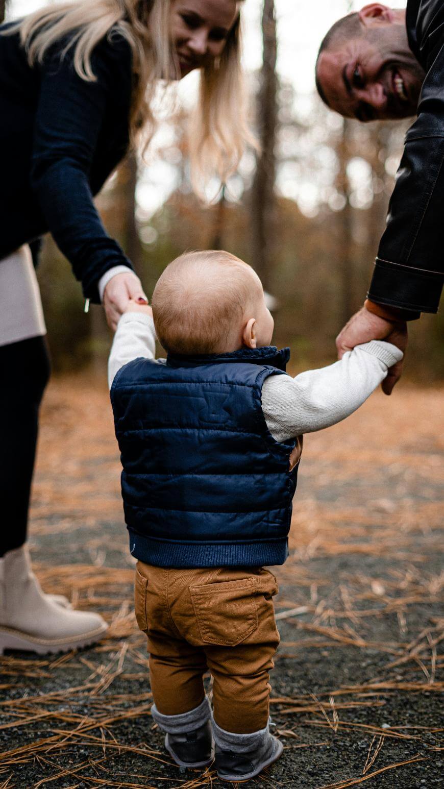 A small child in a navy vest holds hands with two adults, one on each side, while walking outdoors on a pine-covered path. The adults are smiling and looking at the child.