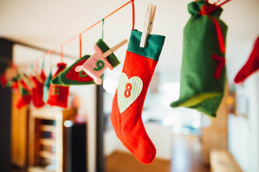 A row of colorful Christmas stockings and mittens, each with numbers and decorations, hangs from a red string with clothespins in a cozy indoor setting. The focus is on a red stocking with a heart and the number 8.