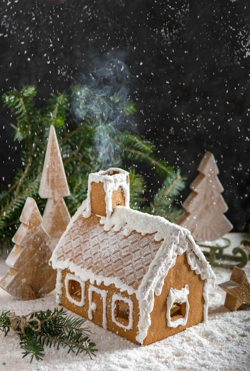 A gingerbread house decorated with white icing sits on a snowy surface. Wooden trees and evergreen branches surround the house, with powdery snow falling and smoke rising gently from the chimney.