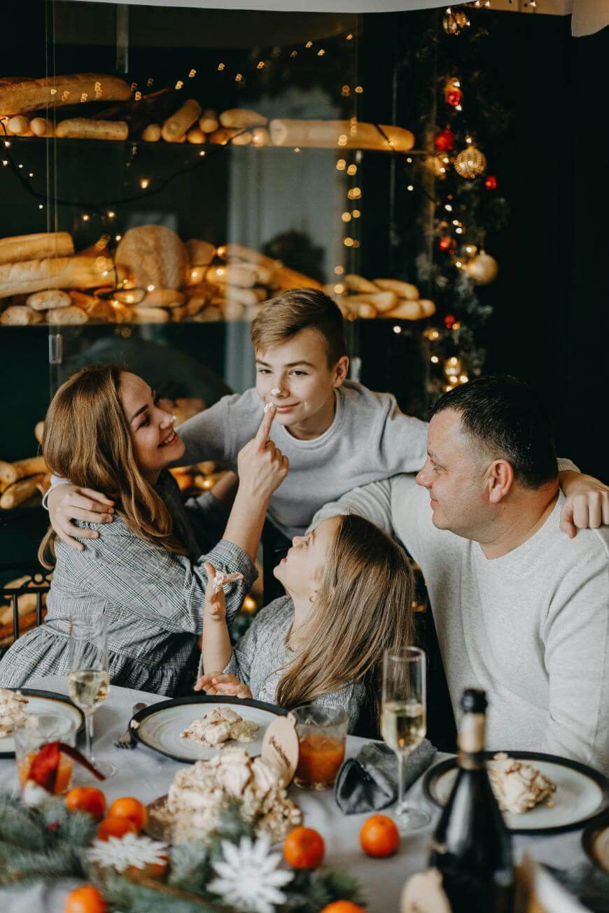A family of four sits at a festive dining table, smiling and interacting warmly. Behind them are shelves with bread and twinkling lights, creating a cozy holiday atmosphere.