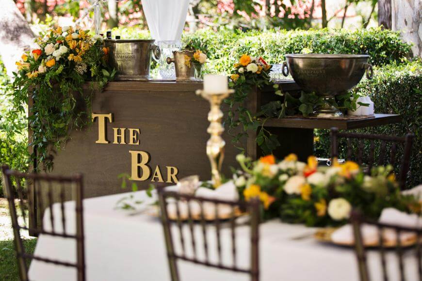 An outdoor bar decorated with lush greenery and orange-yellow flowers, with THE BAR in large gold letters. Nearby, a table is set with floral centerpieces and gold accents. The setting is bright and festive.