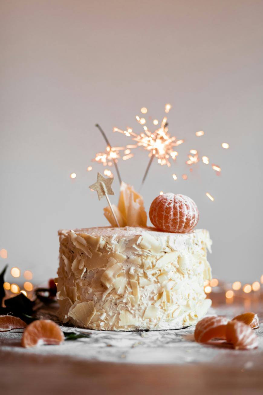 A white chocolate-covered cake topped with a peeled mandarin, decorative stars, and sparklers. The cake sits on a dusted surface with more mandarins around, and blurred lights twinkle in the background.
