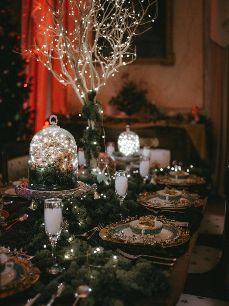 A festive dining table decorated with green garlands, gold-rimmed plates, and glasses of a white beverage, featuring centerpiece branches with twinkling lights and illuminated glass domes creating a warm holiday ambiance.