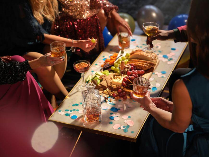 Four people in party attire sit around a table with drinks, a food platter of fruits, cheese, and bread. Balloons and confetti decorate the festive setting.