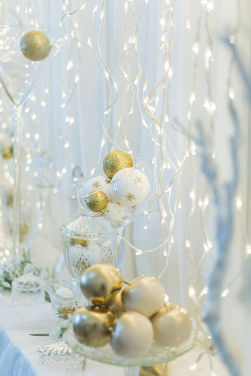 Elegant Christmas ornaments in gold and white are displayed in glass bowls and goblets on a white table, with twinkling string lights and sheer curtains in the background, creating a festive and sophisticated atmosphere.