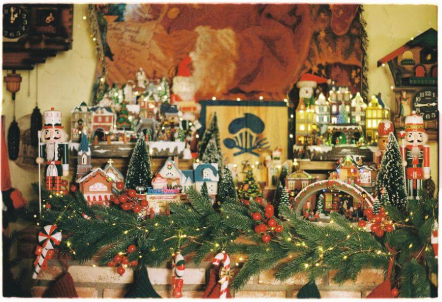 Festive holiday display with miniature Christmas village, nutcrackers, and glowing lights on a decorated mantle with garland, berries, and candy canes in the foreground.