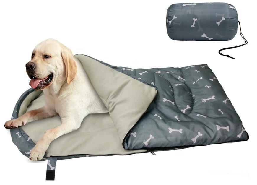 A yellow Labrador retriever lays comfortably inside a gray sleeping bag with a bone pattern. A matching rolled-up sleeping bag is shown above to the right.