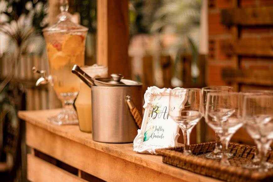 A rustic wooden bar setup with a glass drink dispenser containing citrus-infused water, an ice bucket with tongs, a pitcher, a small framed sign, and empty wine glasses arranged on a tray.
