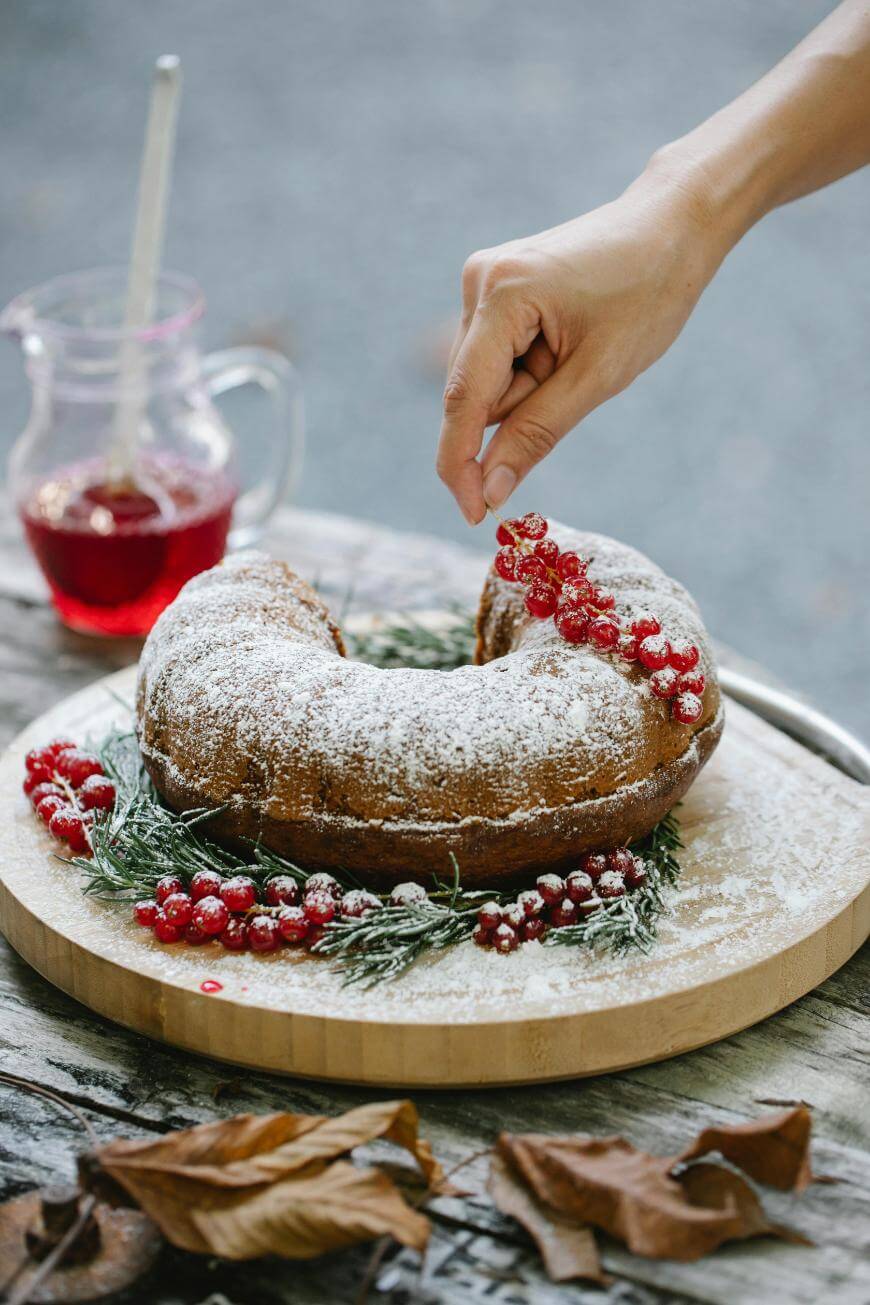 A hand decorates a powdered bundt cake with red berries and greenery on a wooden board, with a glass jug of red liquid in the background and autumn leaves scattered nearby.