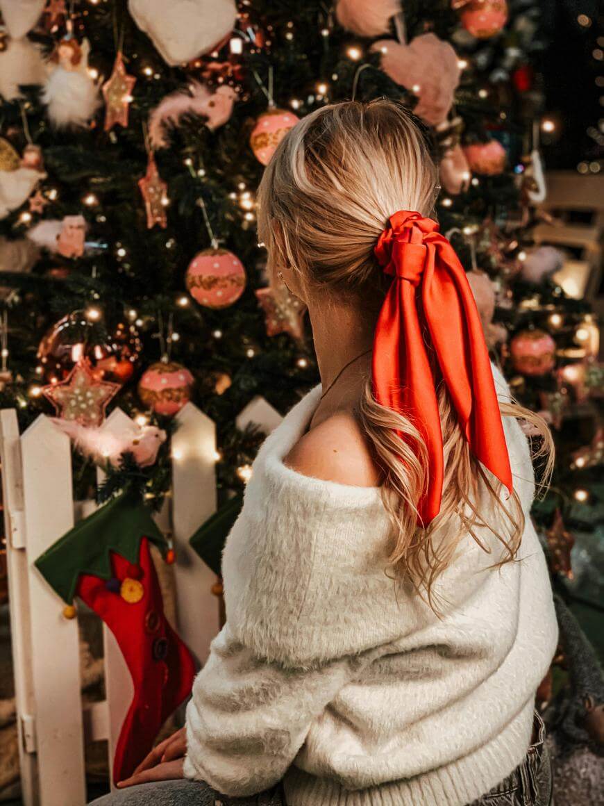 A woman with long blonde hair tied with a red ribbon sits in front of a decorated Christmas tree, wearing an off-the-shoulder white sweater. The scene is festive with ornaments, stockings, and twinkling lights.