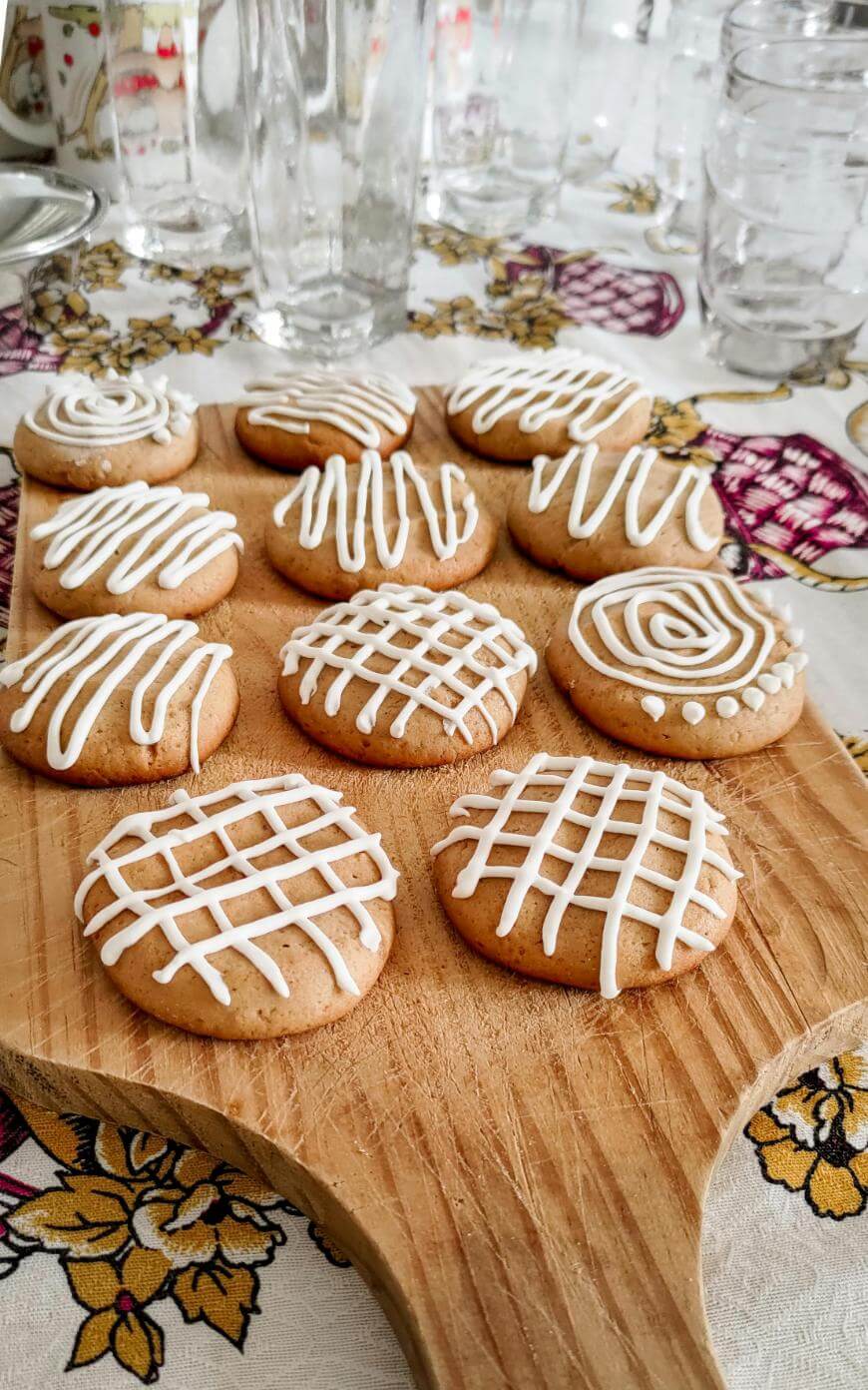 A wooden serving board with round cookies decorated with white icing in various patterns, including swirls, lines, and grids, is set on a patterned tablecloth. Glasses are visible in the background.