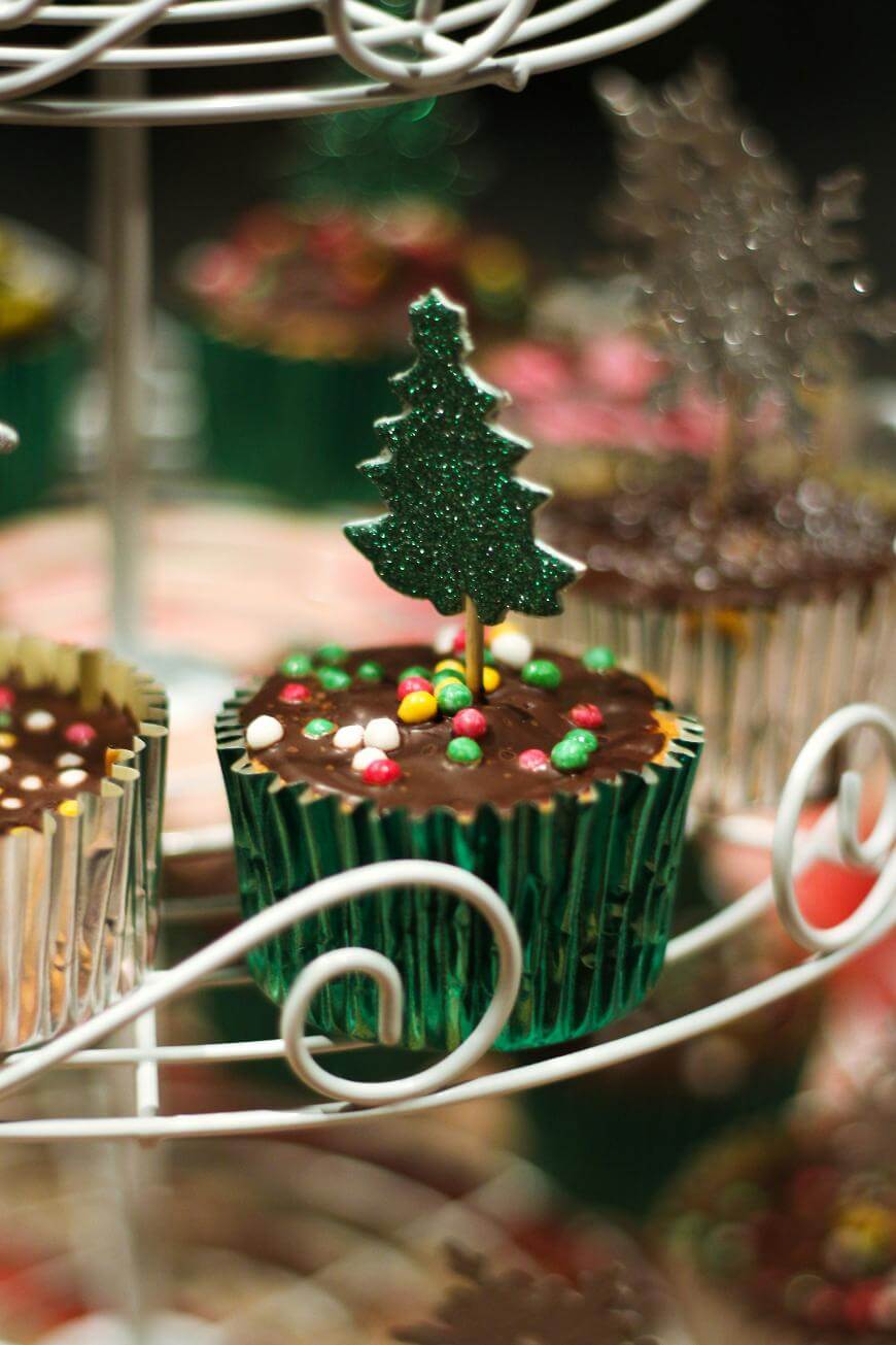 A chocolate cupcake in a green foil wrapper is topped with colorful candy sprinkles and a glittery green Christmas tree decoration, sitting on a metal cupcake stand.