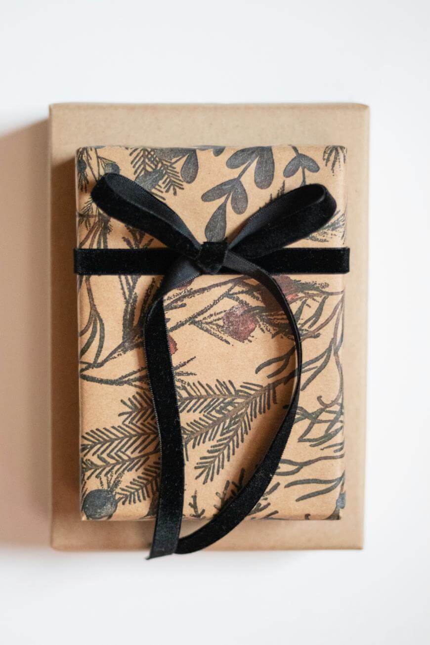 A gift wrapped in brown paper with botanical illustrations, tied with a black velvet ribbon, sits on top of a plain brown box against a white background.