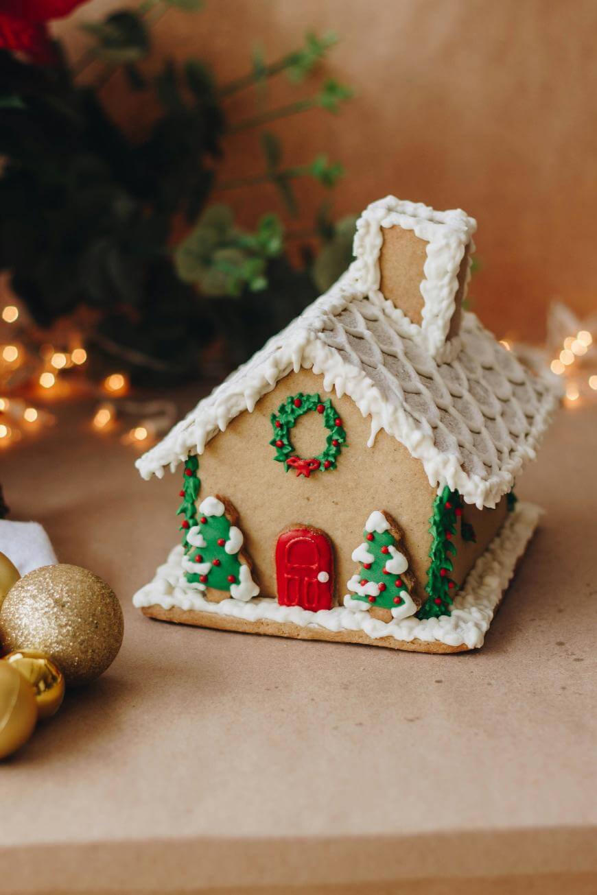 A decorated gingerbread house with white icing, green wreath, red door, and two Christmas trees, sits on a brown surface with gold ornaments and fairy lights in the background.