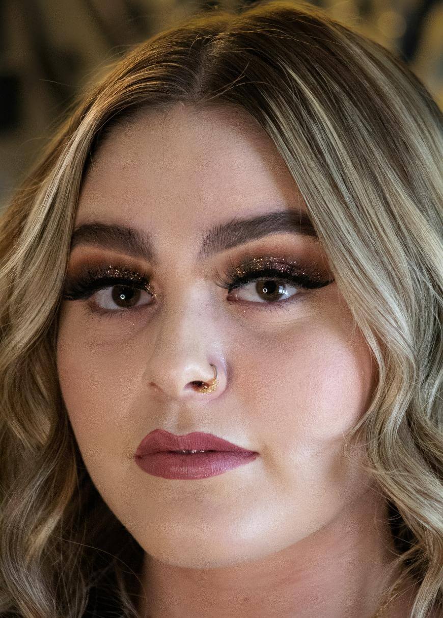 A woman with wavy blonde hair, bold eyebrows, glittery eye makeup, long lashes, and nude lipstick. She has a nose ring and gazes slightly to the side with a calm expression.