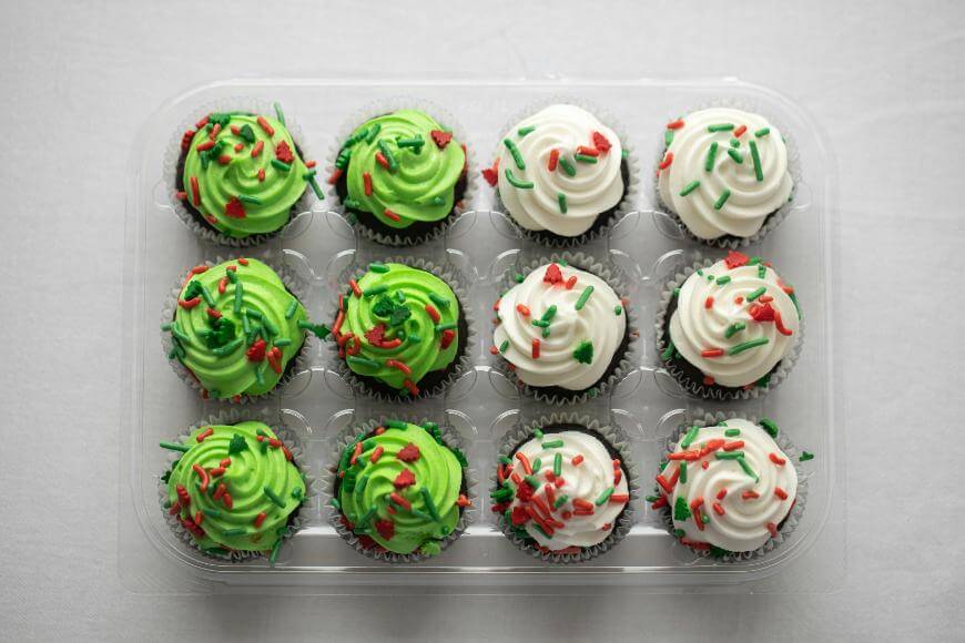 A clear plastic container holds 15 cupcakes, arranged in rows. Half have green icing, half have white icing, all decorated with red and green sprinkles. The container sits on a white surface.