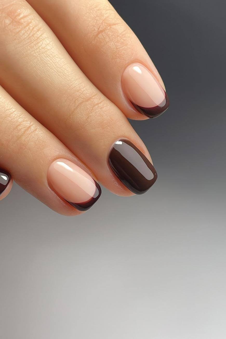 A hand with neatly manicured nails featuring a modern French tip design; the nails are painted nude with thin, dark brown tips, and one nail is fully coated in glossy dark brown polish.