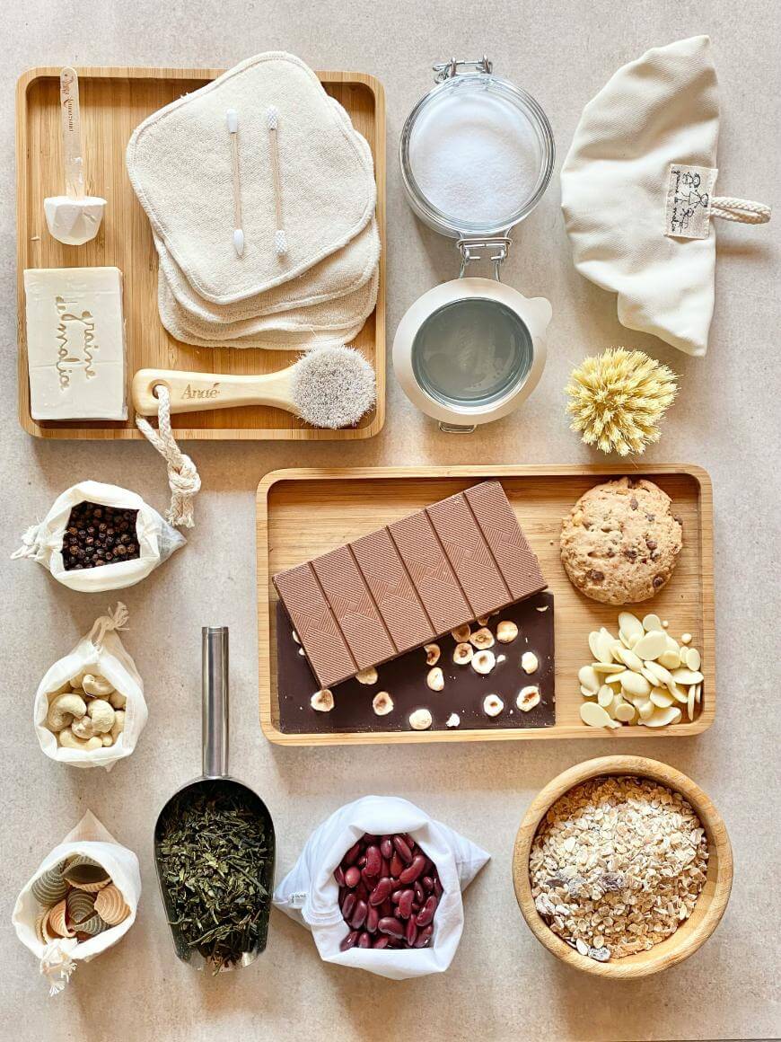 A flat lay of eco-friendly items and foods on a beige surface, including reusable cotton rounds, soap, a brush, cotton swabs, chocolate with nuts, cookies, nuts, spices, grains, beans, a glass jar, and a fabric pouch.