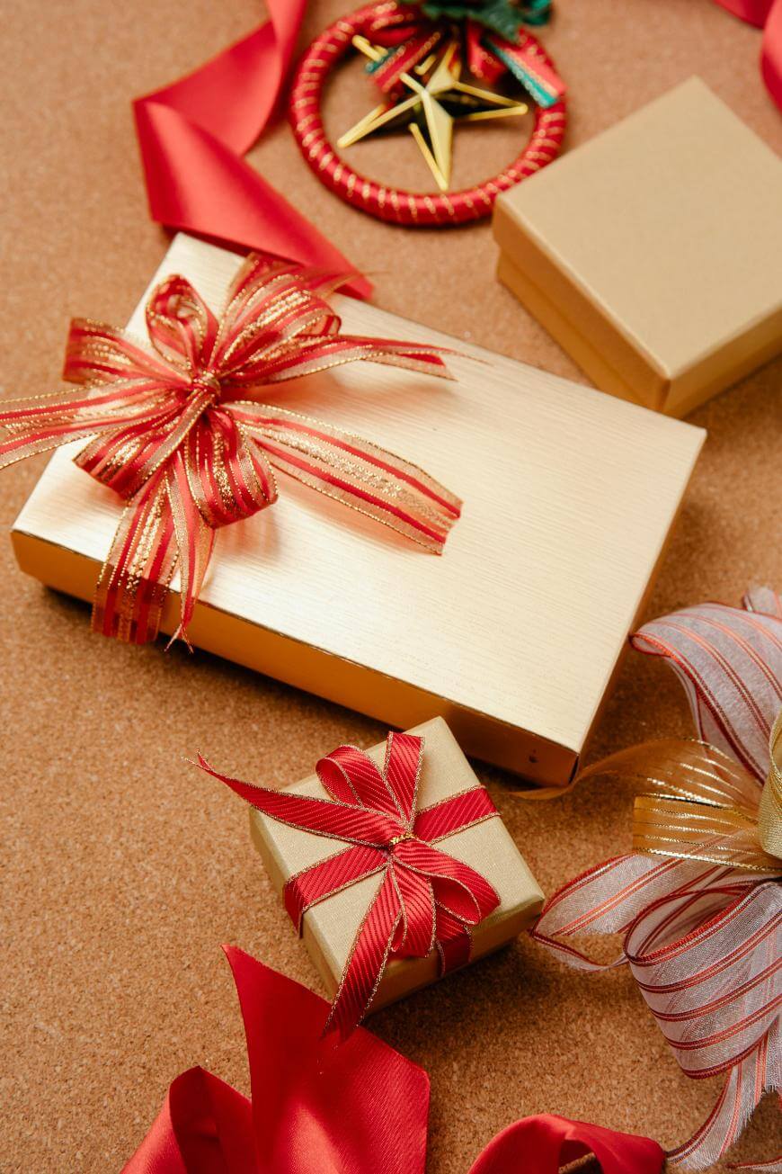 Three gold gift boxes with red and gold bows are arranged on a cork surface, surrounded by red ribbons and a Christmas ornament with a star design.