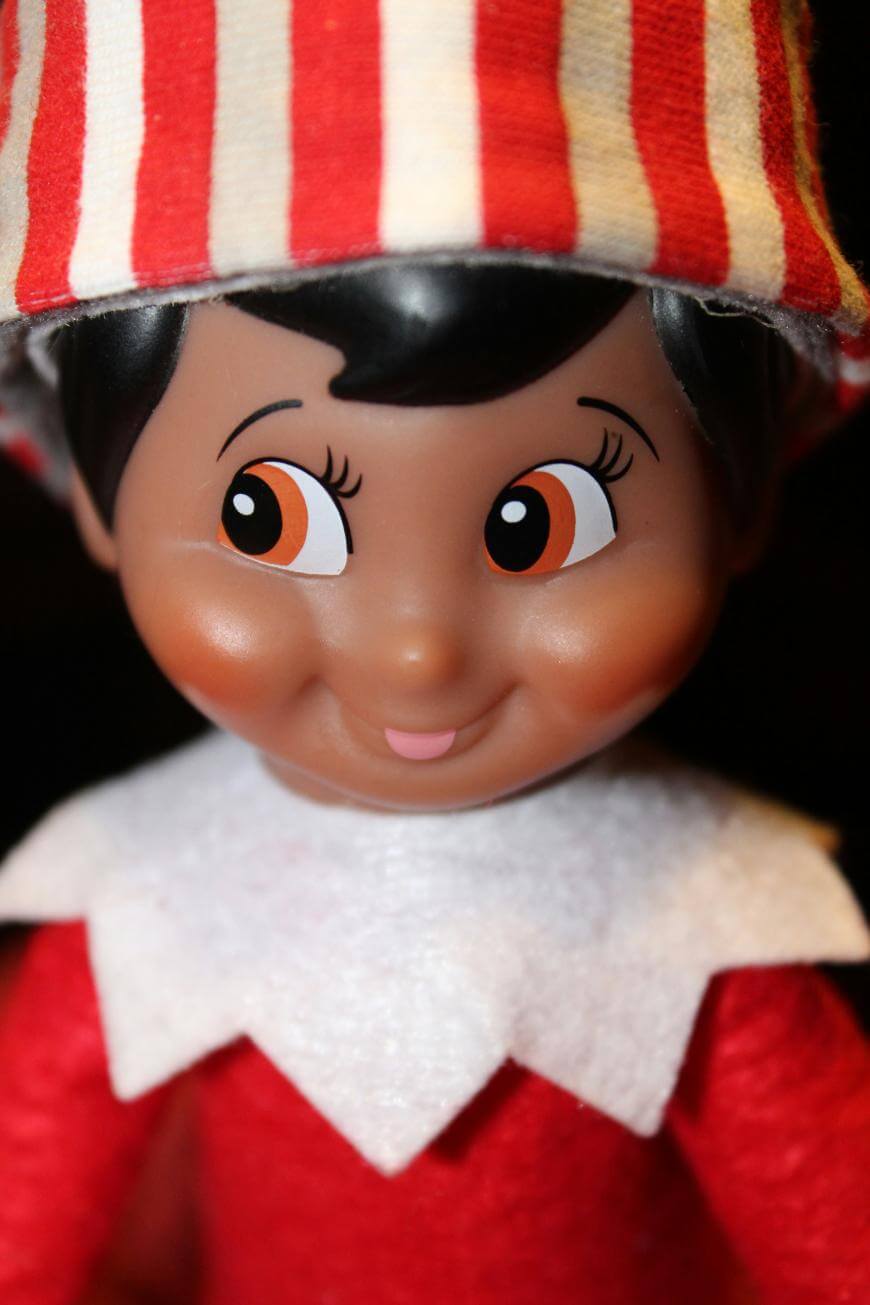 Close-up of a smiling elf doll with brown skin, rosy cheeks, large brown eyes, wearing a red and white striped hat, red outfit, and a white felt collar.