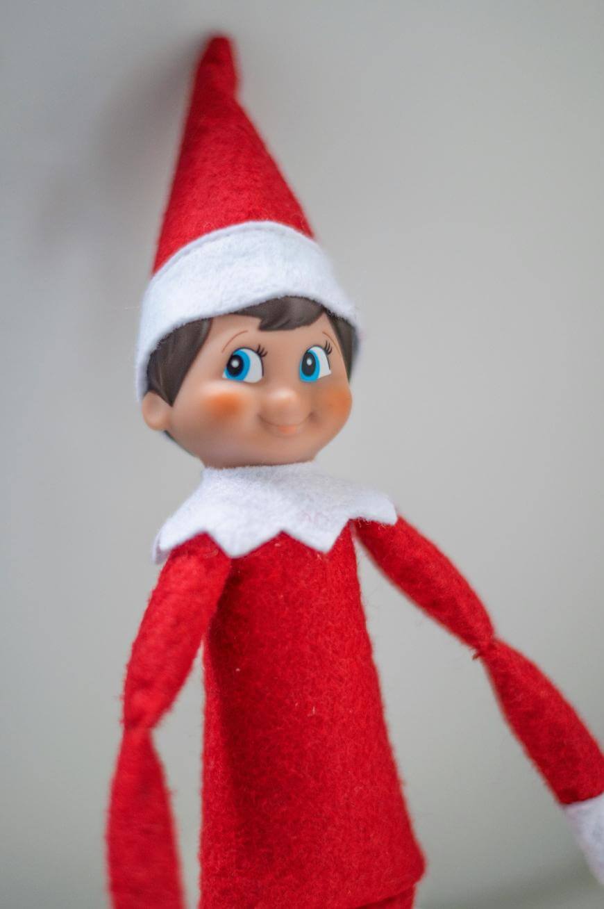 A doll with a red outfit, white collar, and red pointed hat, featuring rosy cheeks, brown hair, and large blue eyes, standing against a plain light background.
