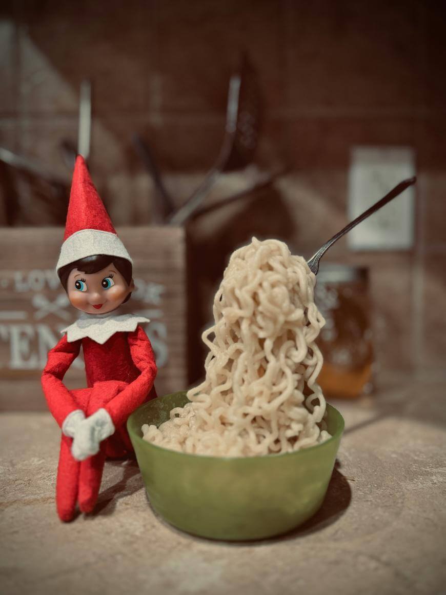 A toy elf in a red outfit sits next to a green bowl filled with noodles, with a fork standing upright in the noodles on a kitchen counter.
