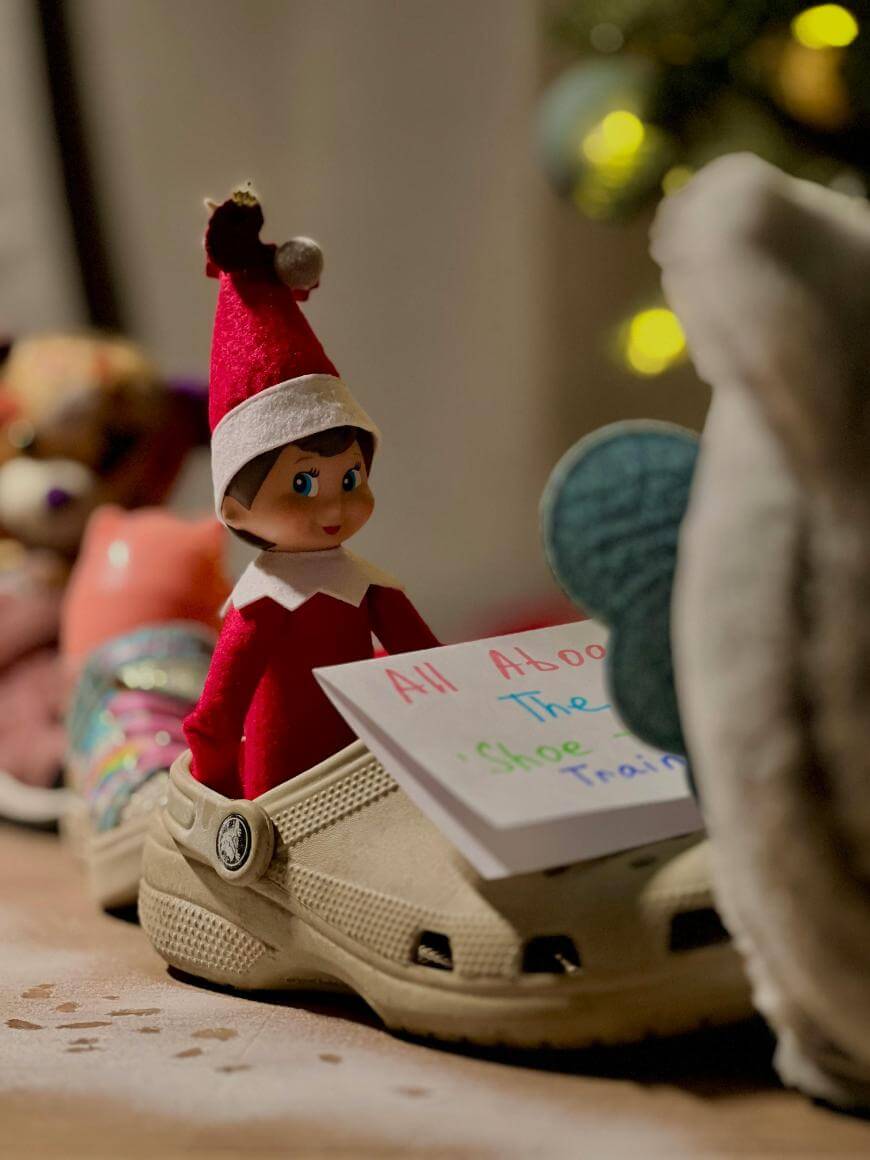 A toy elf in a red outfit sits inside a tan shoe, holding a colorful handwritten sign that says All Aboard the Shoe Train! with other shoes and soft toys in the background.