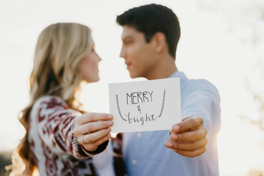 A couple stands close together outdoors, holding a card that says Merry & bright in handwritten script. The focus is on the card, with the couple blurred in the background.