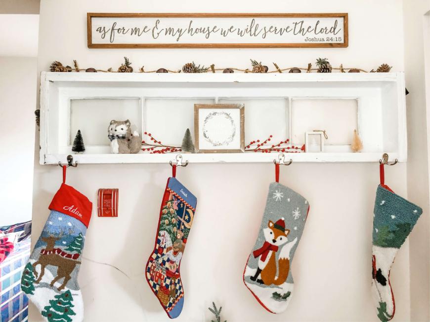 A white shelf with four colorful Christmas stockings hanging below. Decor includes a small wolf figurine, mini trees, and festive garland. Above, a sign reads, As for me & my house we will serve the Lord.