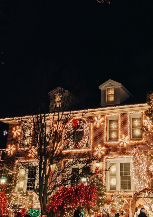 A large brick house is decorated with bright, colorful Christmas lights and ornaments at night. Windows and trees are illuminated, creating a festive holiday atmosphere.