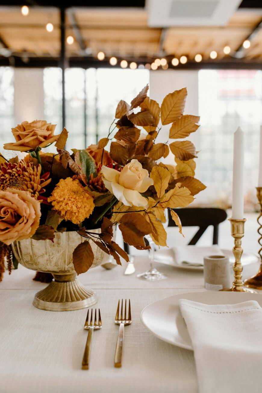Elegant table setting with a white tablecloth, gold utensils, white plate with a cloth napkin, and a centerpiece of yellow and brown flowers. A white candle and gold candlestick are also visible. Warm string lights in the background.