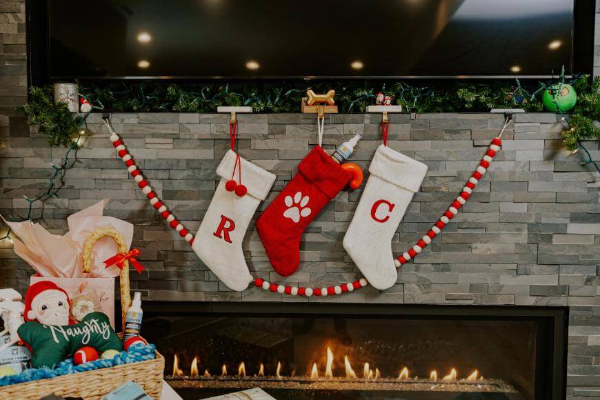 Three Christmas stockings hang on a stone fireplace: two white with red letters R and C, and a red one with a white paw print. Garland and lights decorate the mantel, and a fire burns below. A gift basket sits to the left.