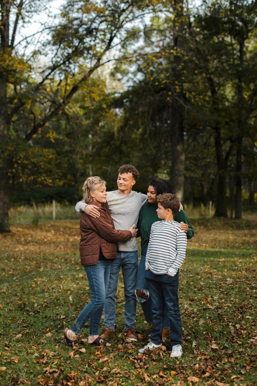 Four people, two adults and two children, stand closely together outdoors in a park with trees and autumn leaves, smiling and hugging each other. The scene looks warm and familial.