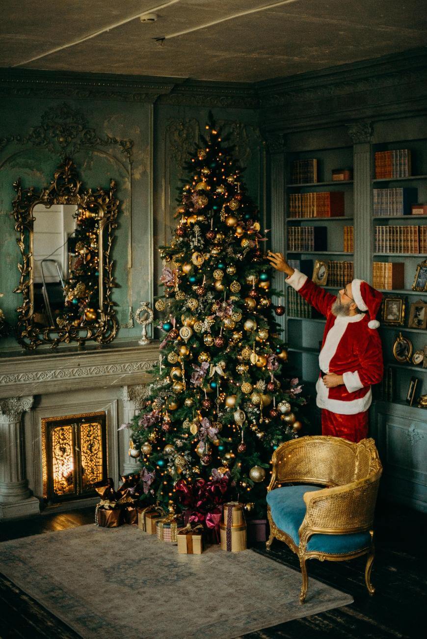 A person dressed as Santa Claus decorates a large, ornate Christmas tree in a vintage-style room with a fireplace, ornate mirror, bookshelves, and wrapped gifts placed under the tree.