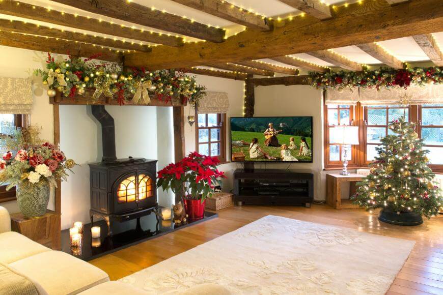A cozy living room decorated for Christmas with garlands, a lit fireplace, poinsettias, a Christmas tree, and a TV showing The Sound of Music, with warm lighting and wooden beams on the ceiling.