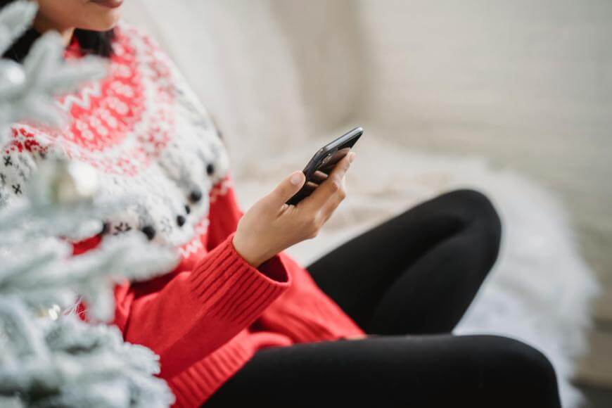 A person in a red and white sweater sits near a frosted Christmas tree, holding a smartphone and looking at the screen. Only the lower part of their face and body are visible.