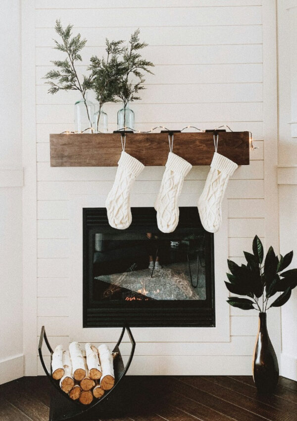 A cozy white shiplap fireplace with a dark wooden mantel, three white knit stockings, glass vases with greenery, a stack of firewood, and a potted plant on the right side.