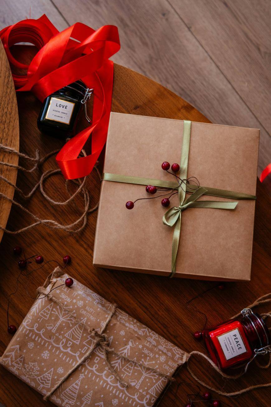 A brown gift box tied with green ribbon and red berries sits on a wooden table, surrounded by red ribbon, twine, two wrapped gifts with festive patterns, and two candles in jars labeled LOVE and PEACE.