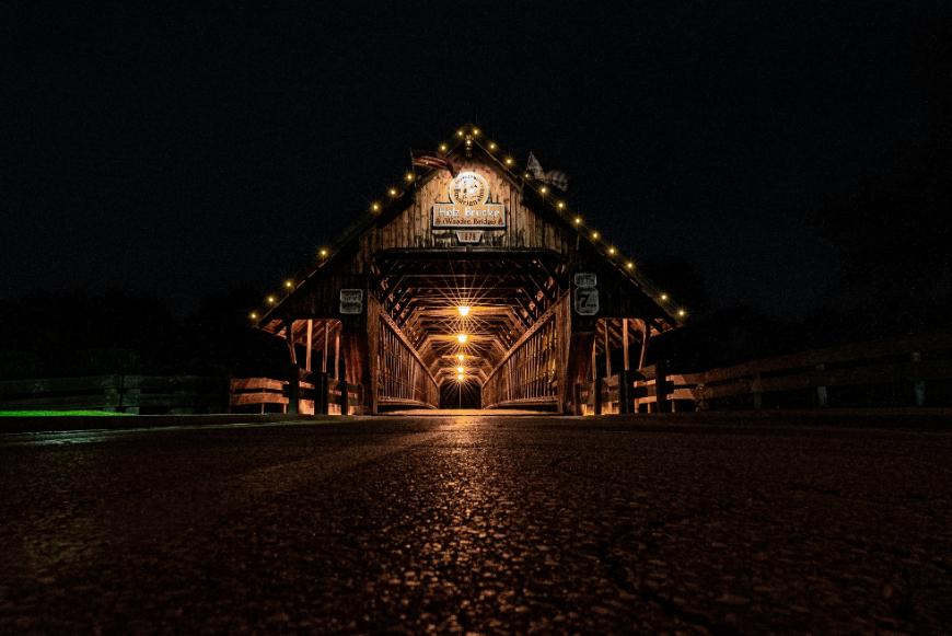 A wooden covered bridge at night is illuminated by warm yellow lights along its roof and interior, casting a glow on the wooden beams and pathway beneath a dark sky.