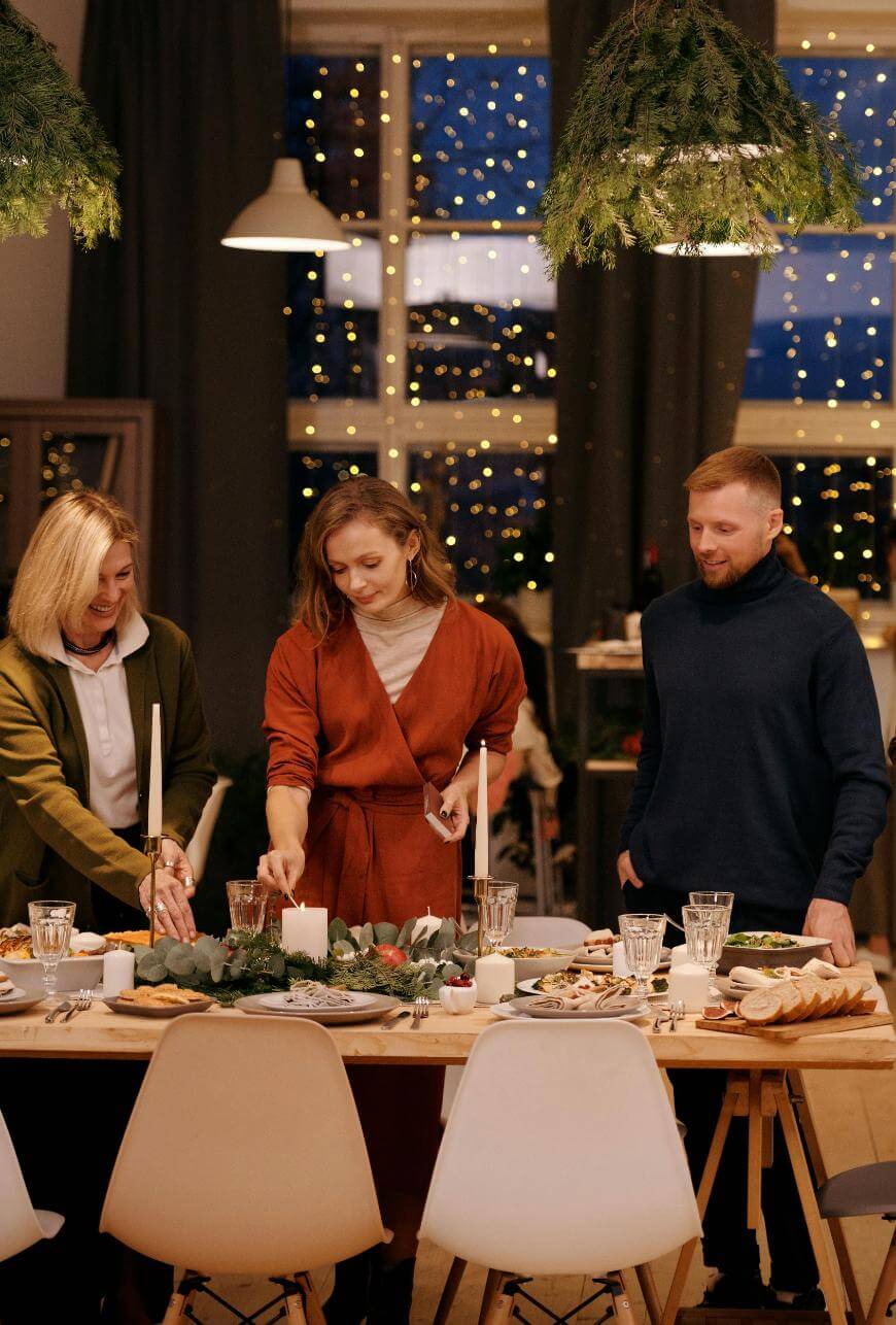 Three people stand around a decorated dinner table with candles, greenery, and food. The room is warmly lit with string lights visible through a large window, creating a festive, cozy atmosphere.