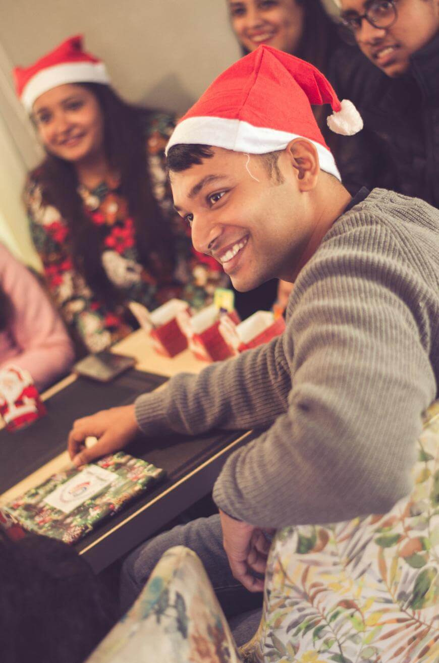 A group of people wearing Santa hats sit around a table, smiling and chatting at a festive holiday gathering. One man in the foreground smiles brightly, while others appear cheerful in the background.
