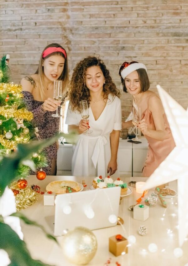 Three women in festive outfits and headbands toast with champagne by a decorated Christmas tree and table, smiling at a laptop during a holiday celebration. Warm lights and ornaments add to the cozy atmosphere.