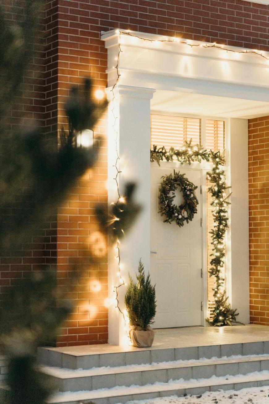 A white front door with a holiday wreath and garland, framed by columns and decorated with warm white string lights. Snow covers the steps, and a small potted evergreen sits by the entrance.