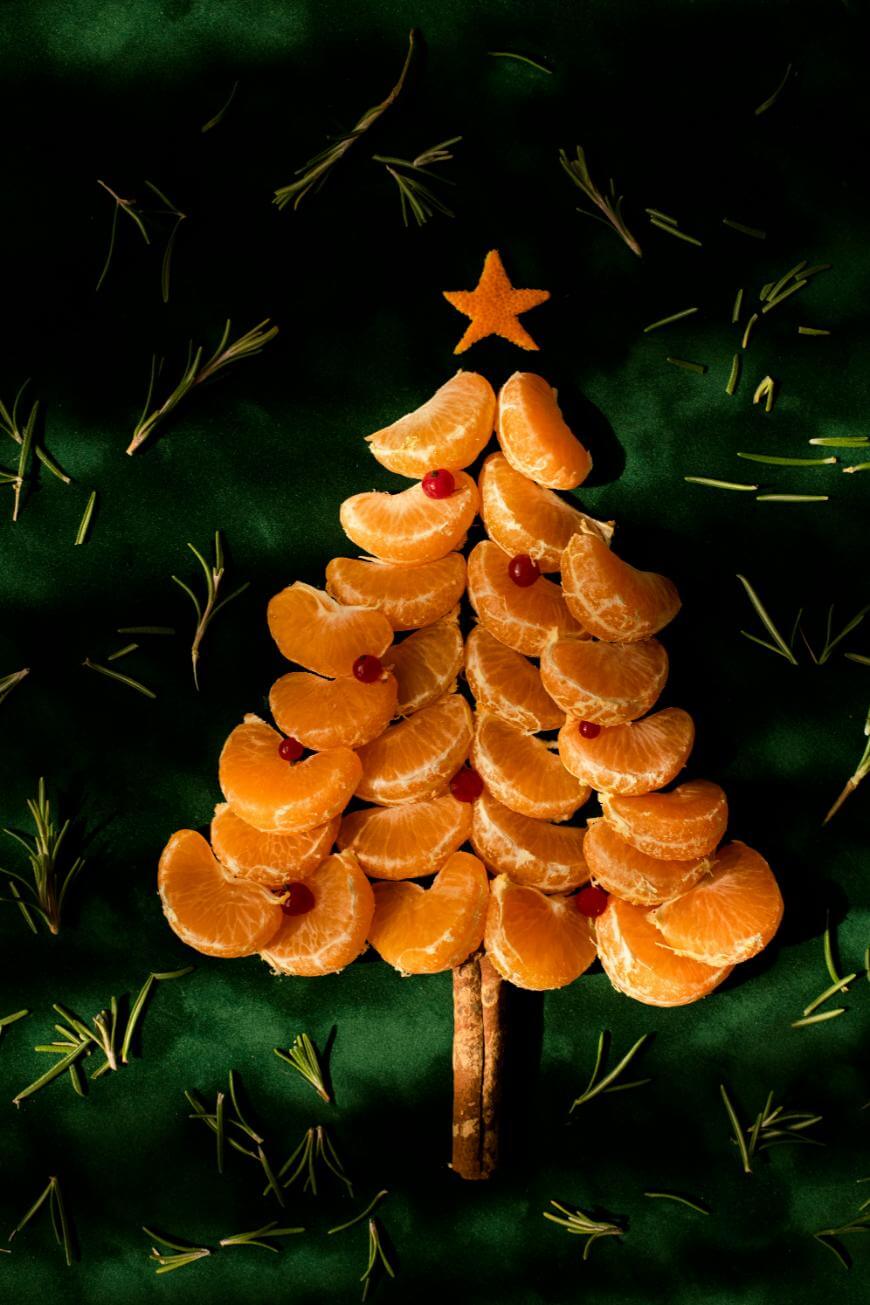A Christmas tree made of tangerine slices, with cinnamon sticks as the trunk, red berries as ornaments, a star-shaped fruit on top, and rosemary sprigs scattered on a dark green background.