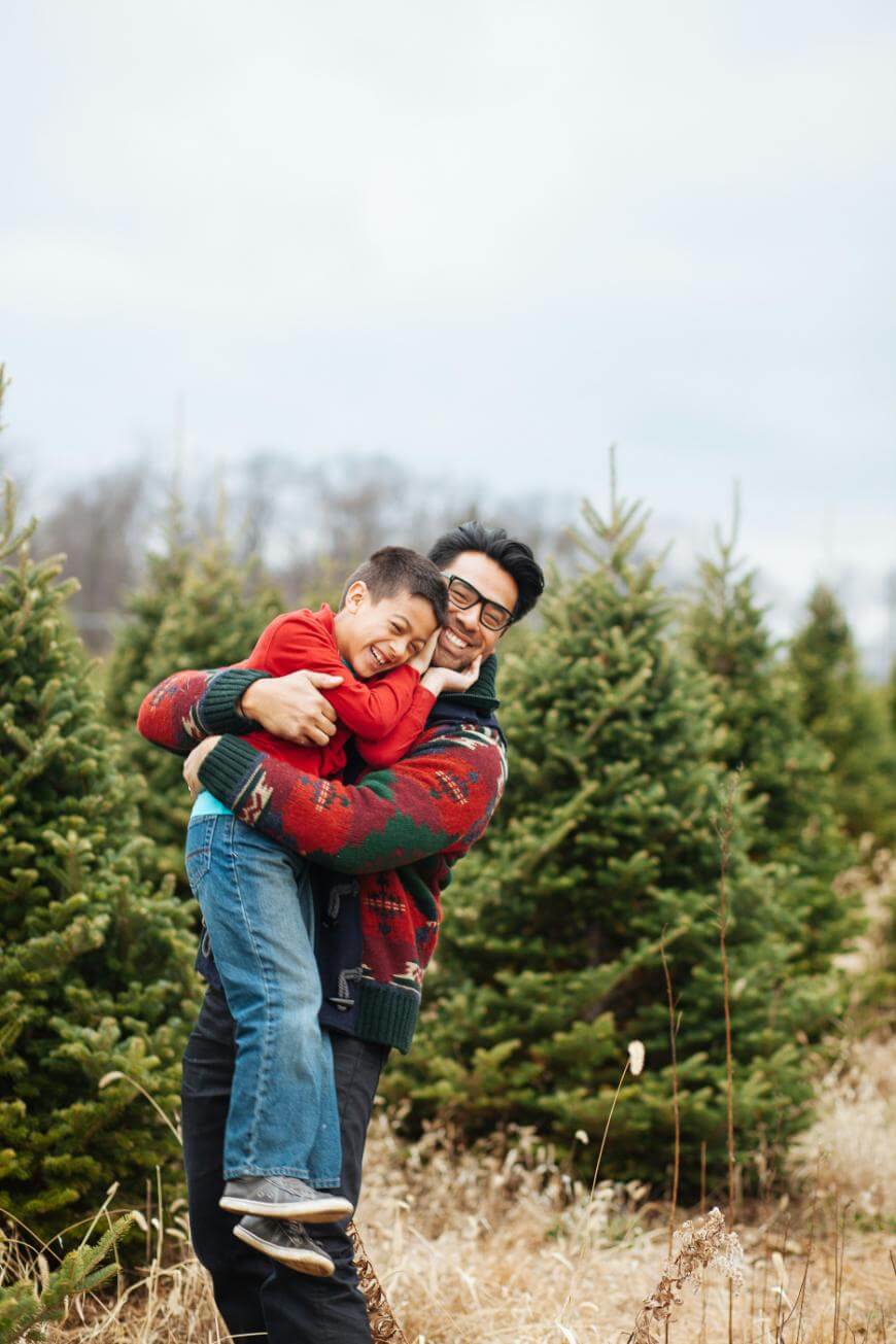 A man wearing glasses and a festive sweater hugs and lifts a smiling young boy in a red shirt among evergreen trees outdoors, both appearing joyful and playful.