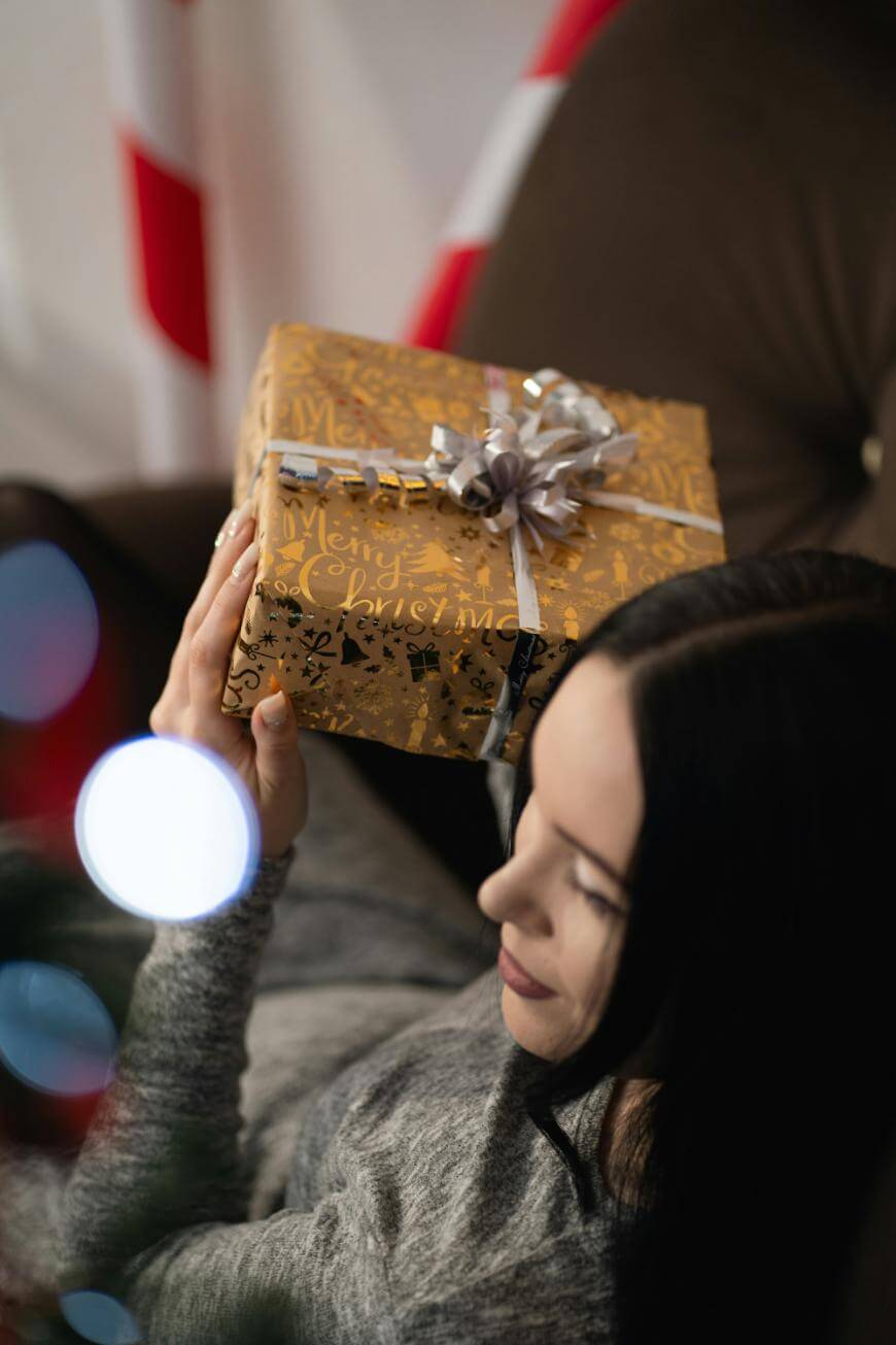 A woman with long dark hair holds a gold-wrapped gift with a silver bow, smiling thoughtfully. She is sitting on a sofa, and the background features blurred holiday decorations.