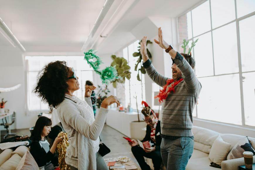 Two people in festive accessories laugh and high-five in a bright, modern office, while others sit and socialize in the background. The atmosphere appears fun and celebratory.
