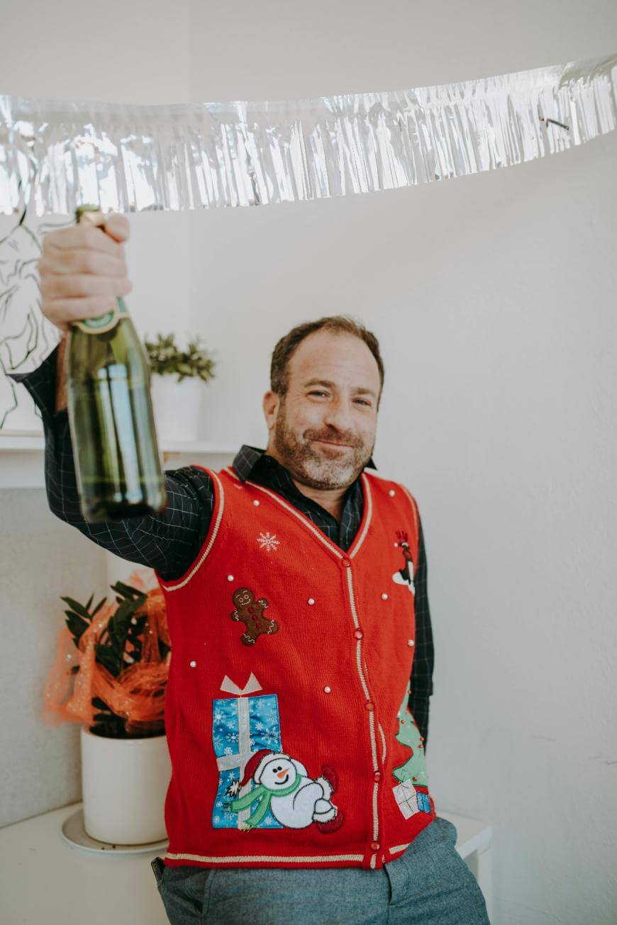 A man in a festive red holiday sweater with winter designs smiles and holds up a bottle of champagne. There is silver tinsel hanging above him and a potted plant wrapped in orange foil in the background.