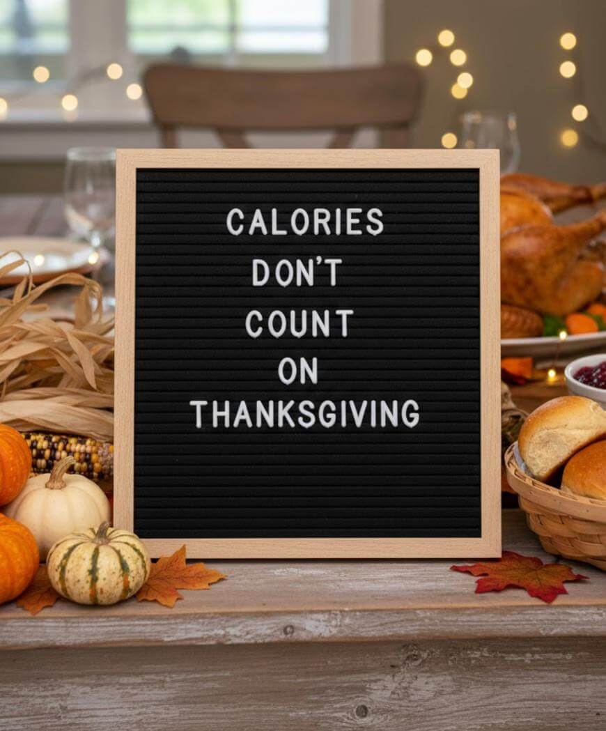 A letter board on a Thanksgiving table reads Calories dont count on Thanksgiving, surrounded by pumpkins, corn, leaves, and festive foods with blurred lights in the background.