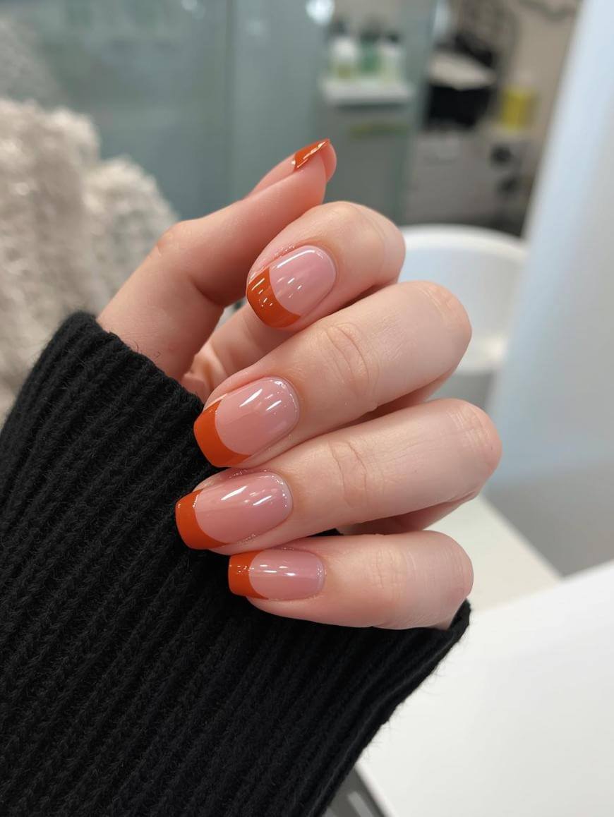 A hand with neatly manicured nails featuring a nude base and bright orange French tips. The person is wearing a black ribbed sweater, and the background is a blurred indoor setting.